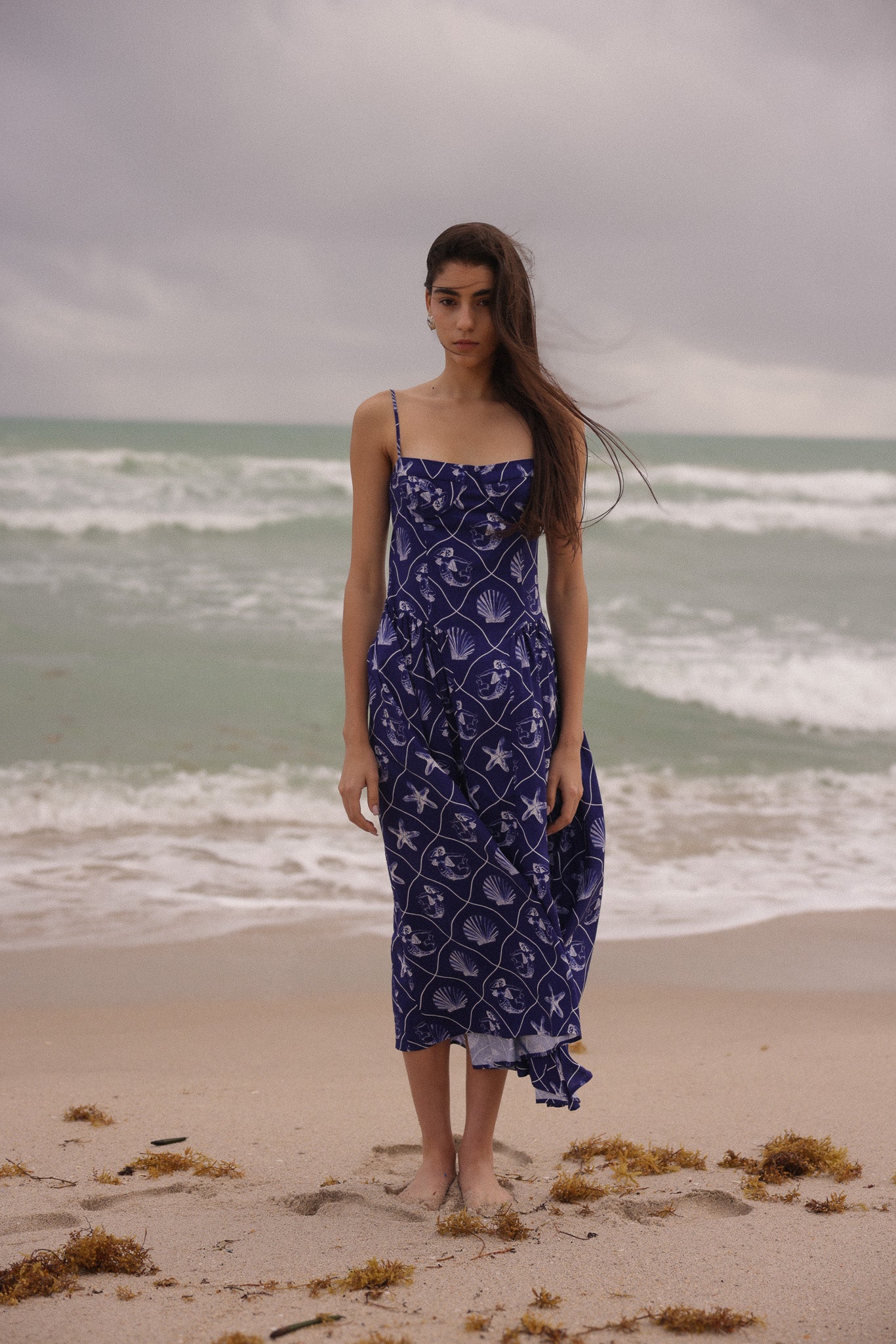 Woman in a blue patterned dress standing on a beach with ocean waves in the background