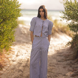 Woman in a light purple outfit standing on a sandy path with greenery and water in the background