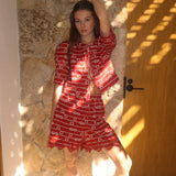 Woman in a red outfit standing against a stone wall with sunlight casting shadows.