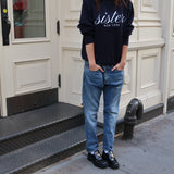 Person wearing a navy hoodie with 'Sister' text, blue jeans, and black shoes standing on a sidewalk.