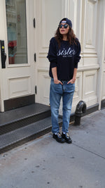 Person wearing a navy hoodie with 'Sister' text, blue jeans, and black shoes standing on a sidewalk.