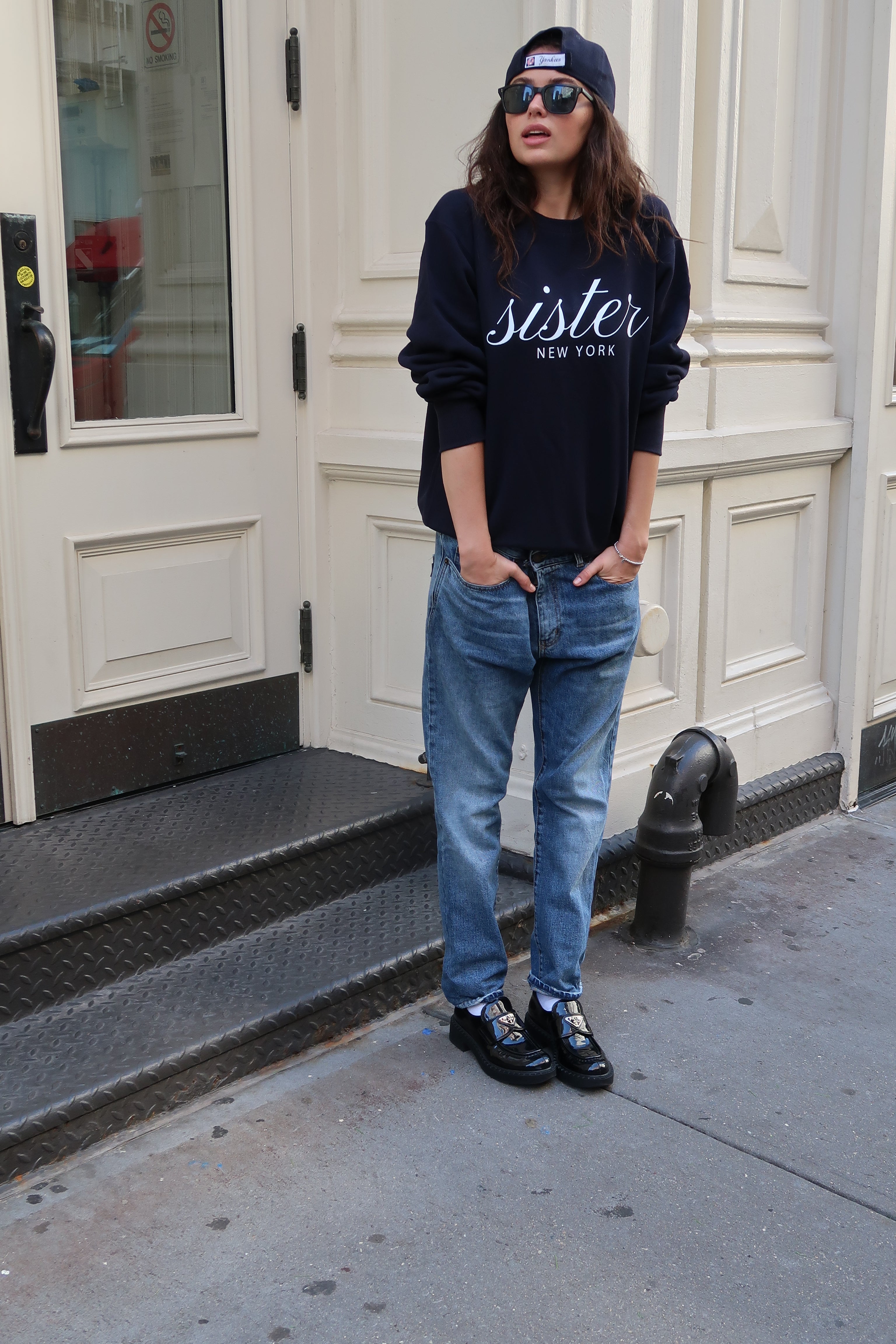 Person wearing a navy hoodie with 'Sister' text, blue jeans, and black shoes standing on a sidewalk.