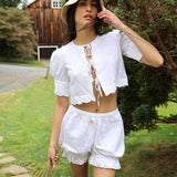 Woman in a white outfit standing outdoors with trees and a wooden building in the background
