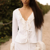 Model walking on a sandy path wearing a white lace-trimmed tie-front top and matching white skirt from Sister NY.