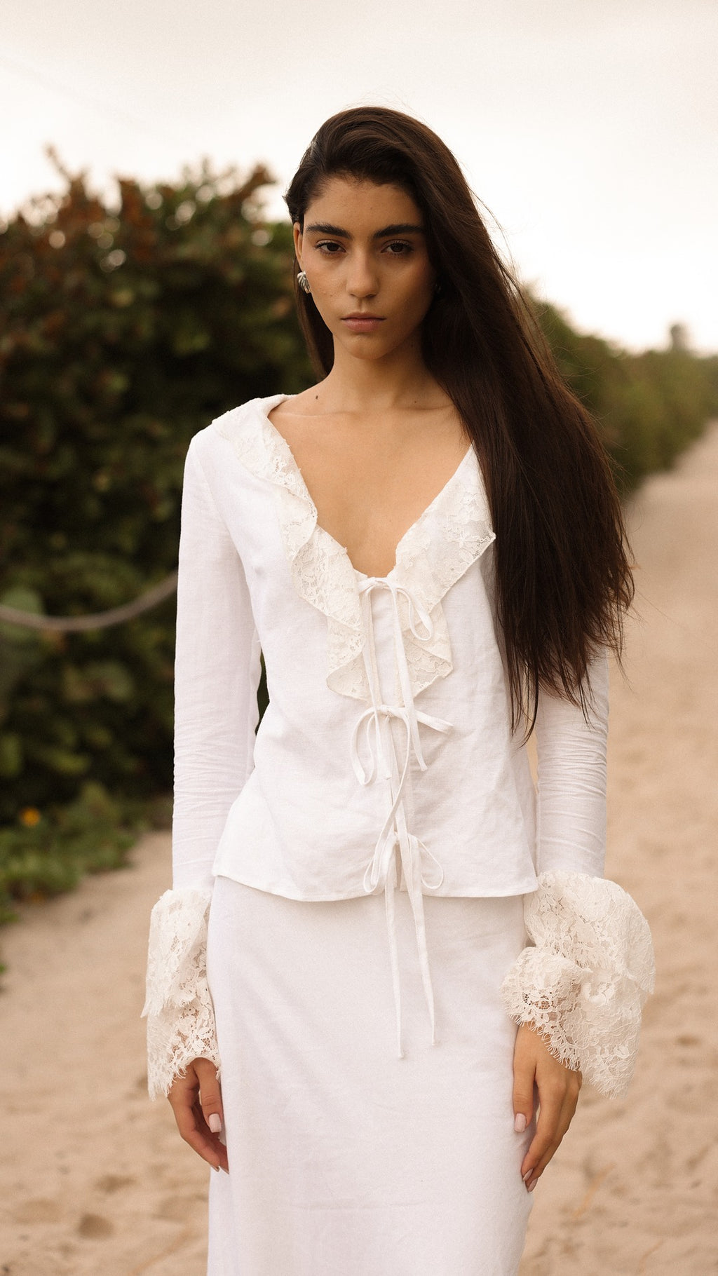 Model walking on a sandy path wearing a white lace-trimmed tie-front top and matching white skirt from Sister NY.