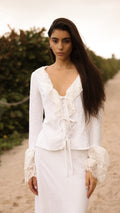 Model walking on a sandy path wearing a white lace-trimmed tie-front top and matching white skirt from Sister NY.