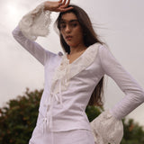 Model wearing a fitted white linen tie-front top with lace trim and dramatic bell sleeves, styled for a romantic summer evening look by Sister New York.