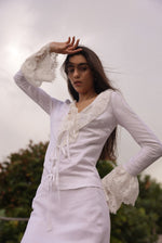 Model wearing a fitted white linen tie-front top with lace trim and dramatic bell sleeves, styled for a romantic summer evening look by Sister New York.
